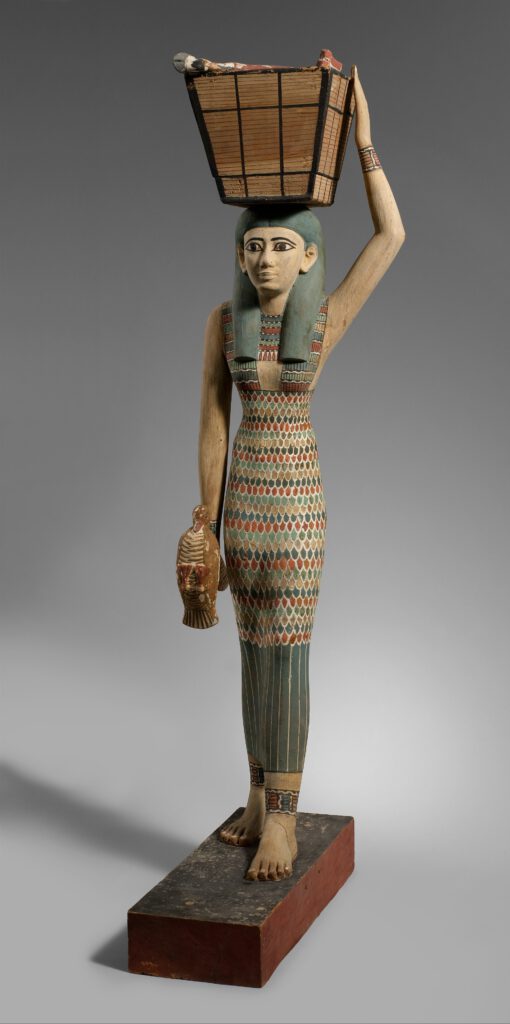 Festgewand. Holzstatue aus Theben um ca. 1900 v. Chr. Auf dem Kopf, Opfergabeen für die Götter, ein Kajal btont die Augen, eine Indigogefärbte Perücke, die Armreifen sind passend zu den Fussreifen, die aus Silber und farbigem Email sind. Das Kleid ist Perlenbesetzt, die Schüre sind aus Leder. 